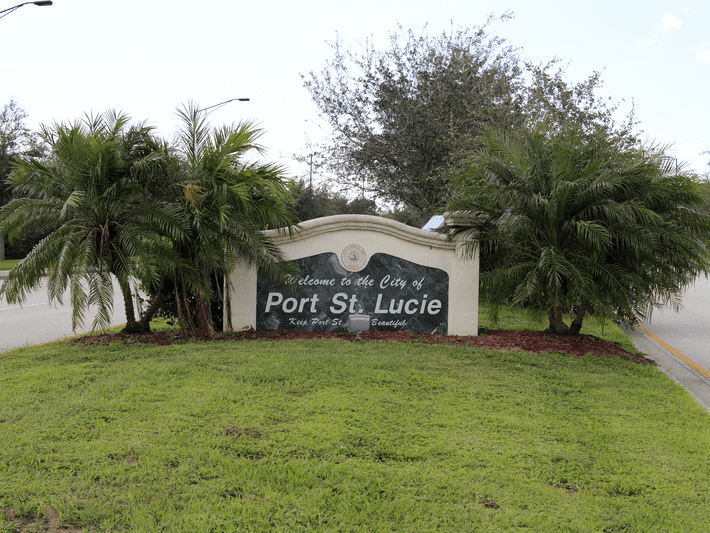 A large sign reading Welcome to the City of Port St. Lucie stands surrounded by palm trees and grass, located at the edge of a roadway.