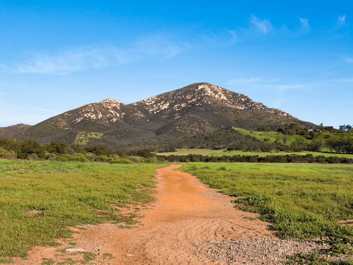 A dirt trail winds through a grassy field toward a mountain with rocky slopes under a clear blue sky.