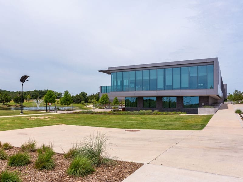 A modern building with large glass windows sits beside a sidewalk, surrounded by green grass, trees, and landscaping. A water fountain is visible in the background under a partly cloudy sky.