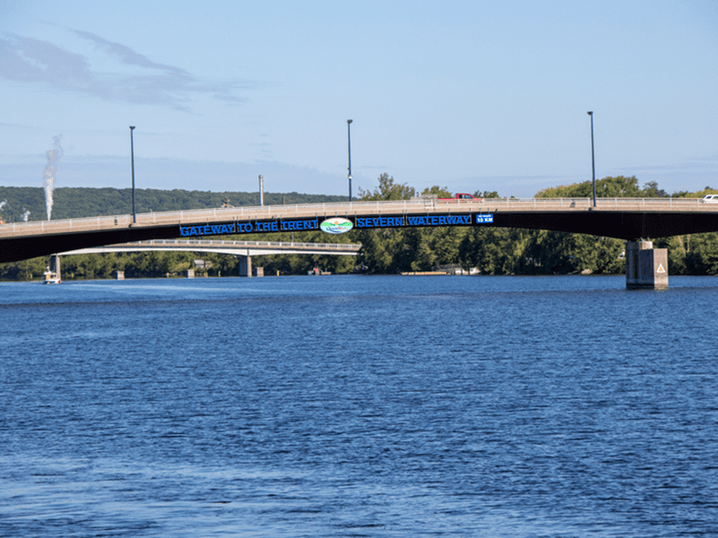 A bridge spans across a wide blue river under a clear sky. The bridge displays a sign reading Gateway to the Trent-Severn Waterway. Trees and a few vehicles are visible in the background.
