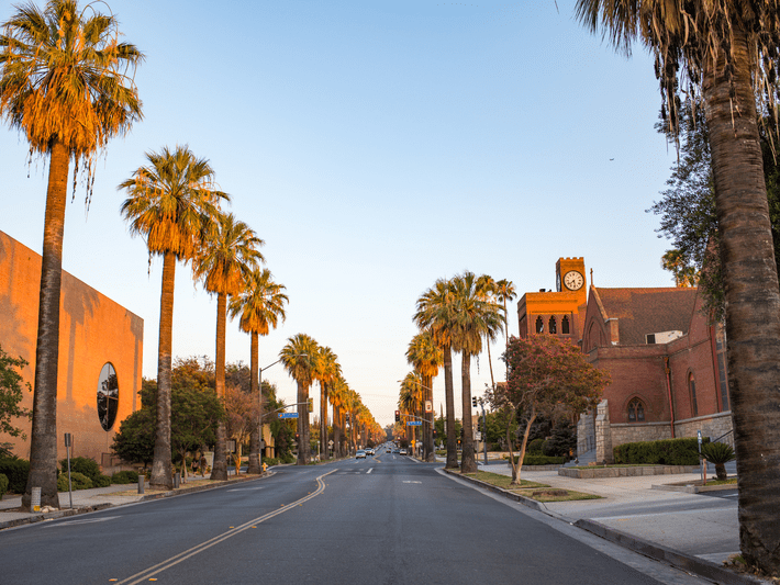 A wide, empty street lined with tall palm trees at sunset, flanked by brick buildings and a red brick church with a clock tower on the right side under a clear blue sky.