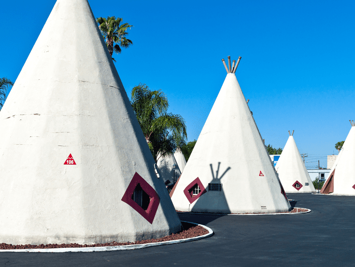 Several white, teepee-shaped motel rooms with red diamond-shaped windows are arranged in a row under a clear blue sky, surrounded by palm trees and an asphalt driveway.
