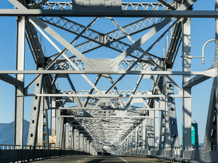 View from a car driving through a steel truss bridge, showing intersecting metal beams and supports against a clear blue sky. Mountains and a road sign are visible in the background.