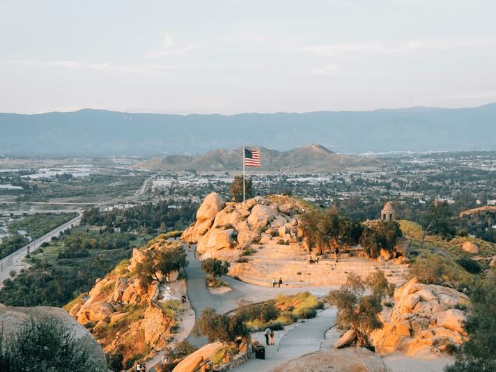 A large American flag atop a rocky hill overlooks a scenic valley, with a winding path, scattered trees, an amphitheater, and distant mountains under a hazy sky.