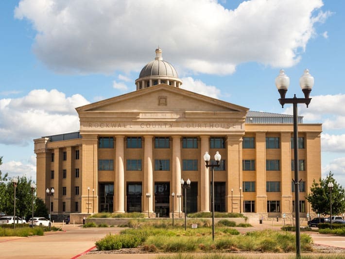 A beige stone courthouse with large columns, arched windows, and a dome on top, labeled Rockwall County Courthouse, under a blue sky with scattered clouds. Lamp posts and landscaping are in front.