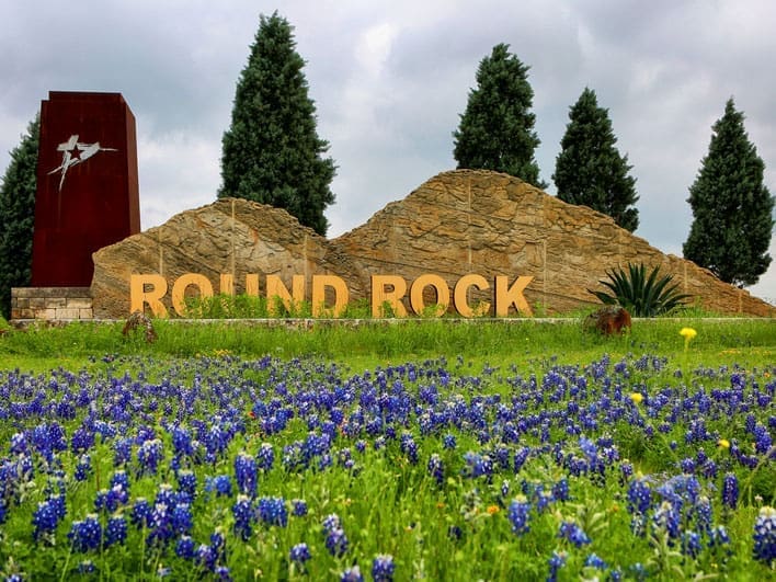 A large stone sign with yellow letters reads ROUND ROCK, surrounded by blooming bluebonnet flowers and tall trees under a cloudy sky.