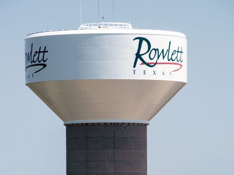 A large white water tower with Rowlett Texas written on the side in blue and red script, set against a clear blue sky.
