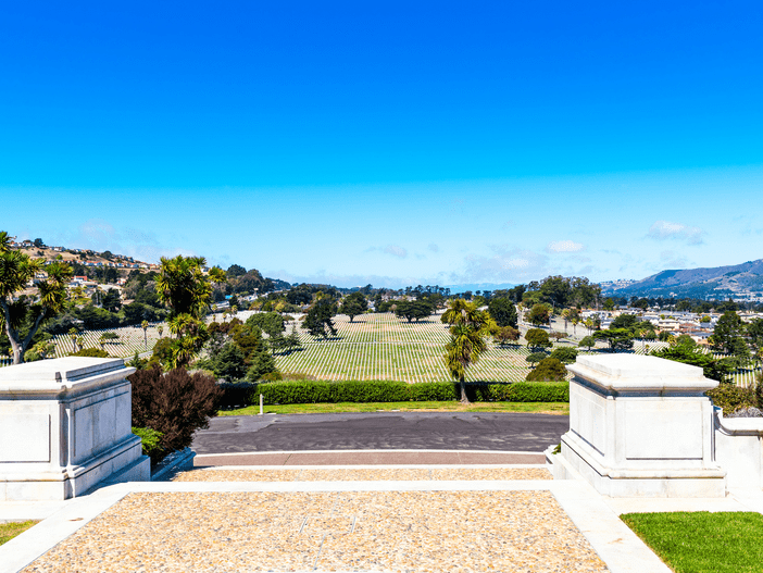 View from a stone terrace overlooking a cemetery with rows of headstones, palm trees, grassy lawns, and distant hills under a clear blue sky.