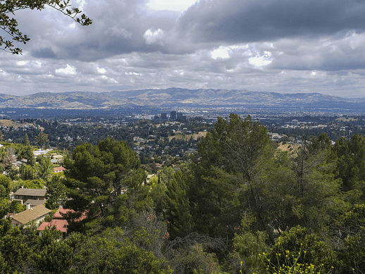 A scenic view of a suburban area with scattered houses, dense green trees in the foreground, and distant mountains under a cloudy sky.