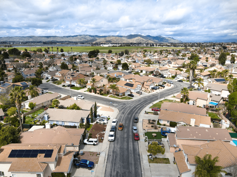 Aerial view of a suburban neighborhood with rows of tan-roofed houses, parked cars lining the streets, palm trees scattered throughout, and mountains visible in the background under a partly cloudy sky.