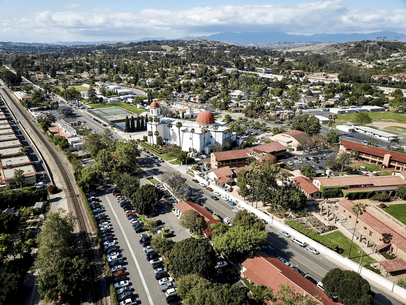 Aerial view of a mission-style building with a red dome and white walls, surrounded by trees, parking lots, railroad tracks, and a suburban neighborhood, with hills and mountains in the distance.