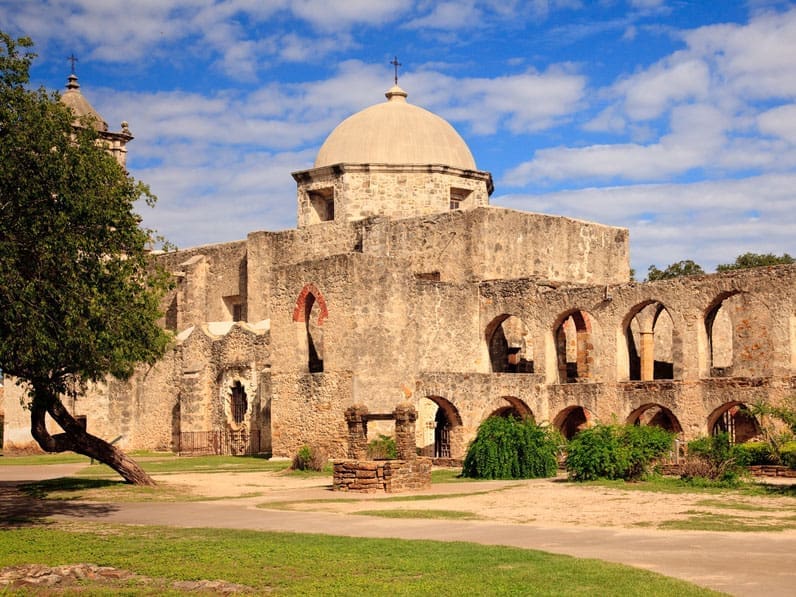 Historic stone mission building with arched walkways, a central dome, and a small cross on top, surrounded by green trees and bushes under a blue sky with scattered clouds.