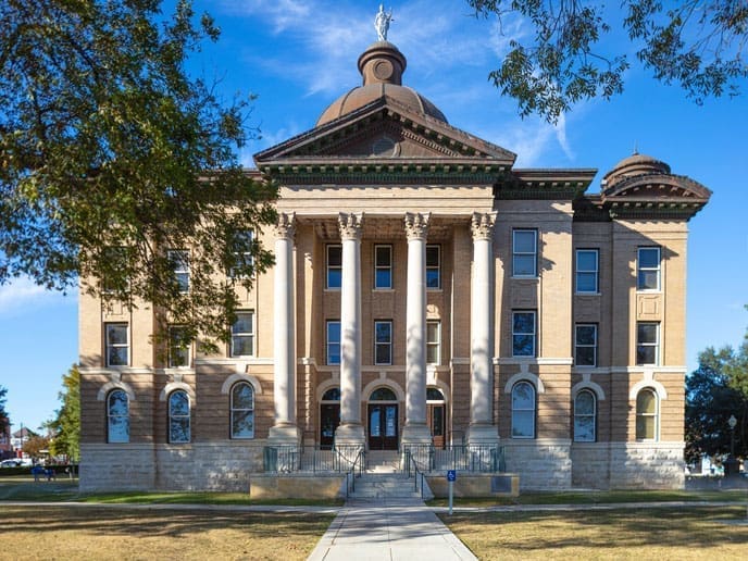 A historic courthouse with large columns, arched windows, and a domed roof stands on a grassy lawn under a blue sky, framed by tree branches.