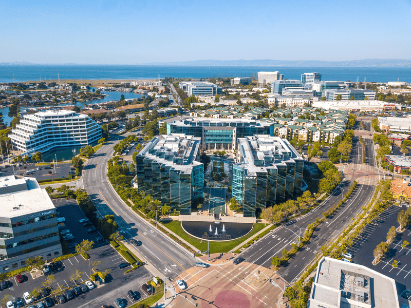Aerial view of modern office buildings with glass facades, arranged in a circular pattern near a main road, surrounded by parking lots, trees, and other commercial buildings, with water and cityscape in the background.
