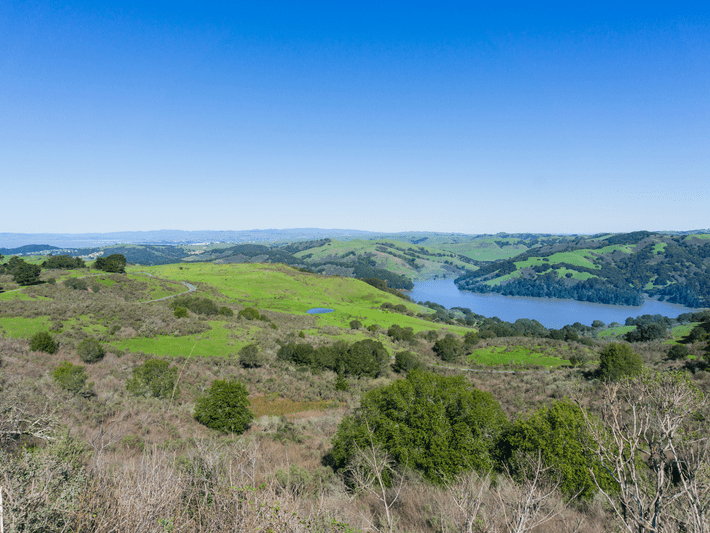 A scenic landscape with rolling green hills, scattered trees, and a large blue lake under a clear, bright blue sky. The view extends into the distance, showcasing a peaceful, natural environment.