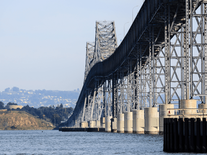 A long steel bridge stretches across the water with large support pillars underneath. Distant hills and greenery are visible in the background under a clear sky.