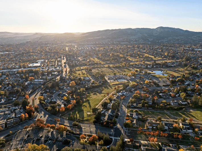 Aerial view of a suburban neighborhood with houses, roads, trees with autumn foliage, and distant hills under a clear sky at sunset.