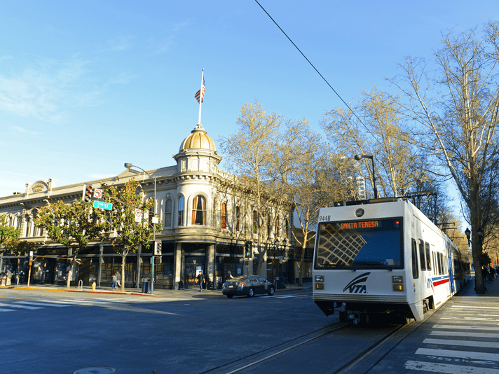 A white VTA light rail train travels through a sunny downtown street with historic buildings, leafless trees, cars, and a clear blue sky in the background.