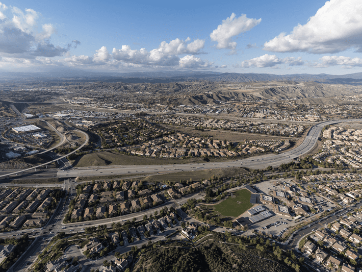 Aerial view of a suburban area with houses, roads, and highways, surrounded by hills and mountains under a partly cloudy sky. Green spaces and a sports field are visible in the foreground.