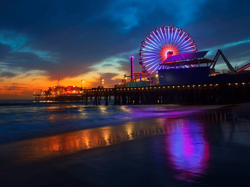 A brightly lit Ferris wheel and amusement park glow with colorful lights on a pier at sunset, reflecting on the wet sand and ocean waves beneath a dramatic blue and orange sky.