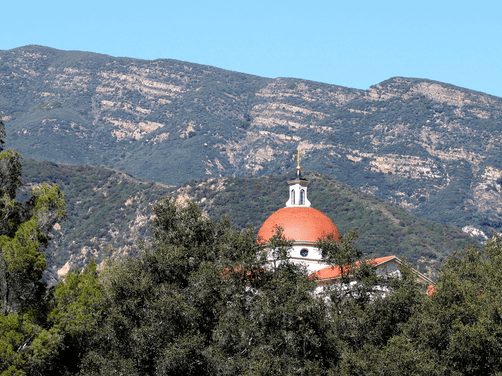 A red-domed building partially hidden by trees, set against green mountains with layered rock formations under a clear blue sky.
