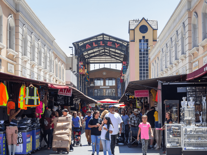 A busy outdoor market alley with people walking, clothing and jewelry stalls on both sides, and a large sign reading “ALLEY” overhead on a sunny day.