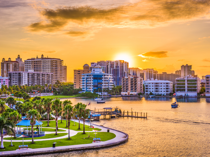 A vibrant sunset over a waterfront cityscape with modern high-rise buildings, palm trees along a park, boats on the water, and a golden sky reflecting off the river.