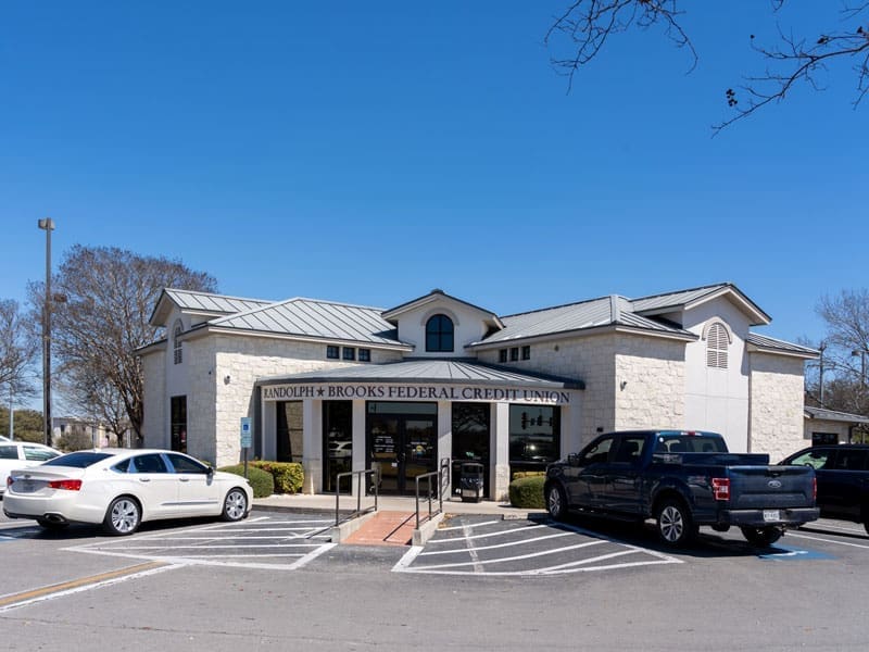 Single-story Randolph-Brooks Federal Credit Union building with stone exterior, large front windows, and two accessibility parking spaces; several cars are parked in front under a clear blue sky.
