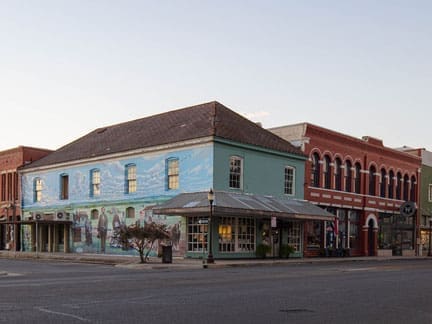 A corner building with a mural painted on its blue side wall stands at an empty intersection, next to other historic brick buildings under a clear sky.