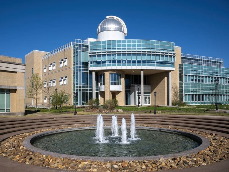 A modern academic building with large glass windows and an observatory dome on top, stands behind a circular fountain with four water jets, surrounded by landscaped stonework under a clear blue sky.