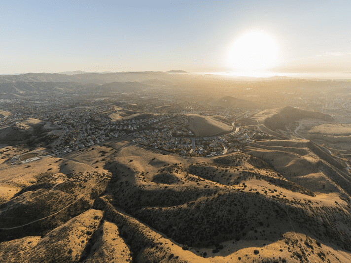 Aerial view of a sunlit landscape with rolling hills, scattered vegetation, and a sprawling town below, taken at sunrise or sunset with the sun low on the horizon and distant mountains in the background.