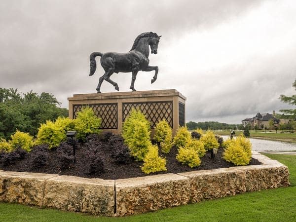 A bronze horse statue stands on a stone pedestal surrounded by yellow-green shrubs and a rock border, with overcast skies and a pond in the background.
