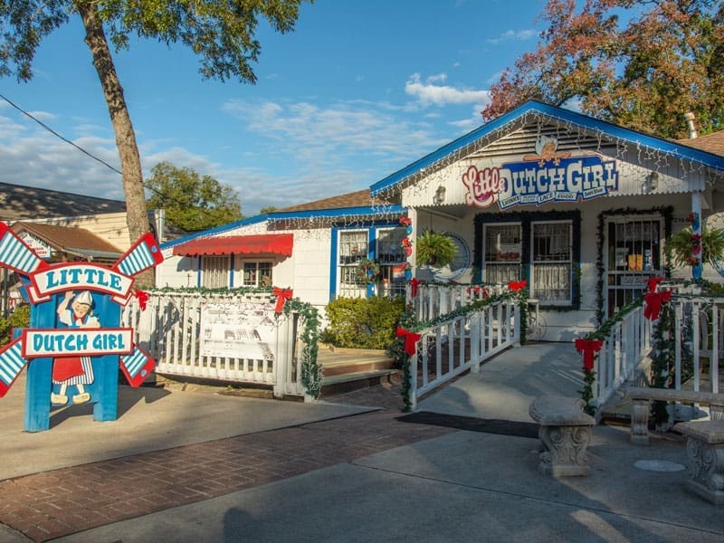 A quaint shop called Little Dutch Girl decorated with red bows, garlands, and a festive sign, with a white porch and a wooden ramp under a blue sky with scattered clouds.