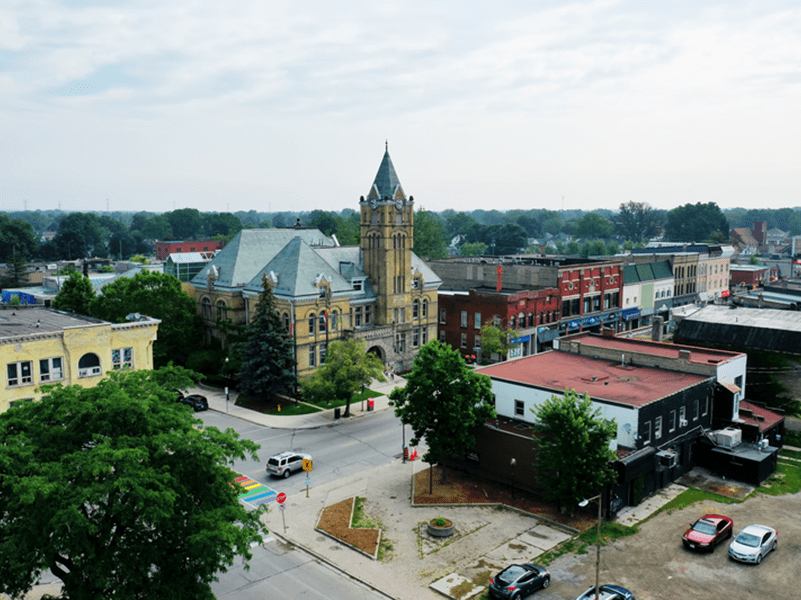 Aerial view of a small town with a historic stone building featuring a clock tower, surrounded by trees, parked cars, and rows of brick storefronts along the streets.