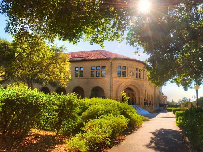 A sunlit, historic building with arched windows and a red-tiled roof is partially shaded by large green trees, with a pathway and manicured bushes in the foreground.