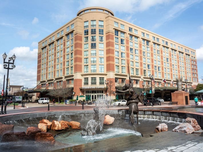 A large hotel building with a Starbucks on the ground floor stands behind a circular fountain featuring a bronze statue of a horse and rider in a city plaza on a sunny day. Cars and street lamps are visible nearby.
