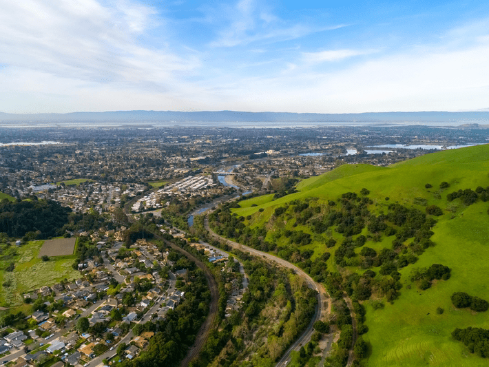 Aerial view of a suburban neighborhood next to green hills, with a winding river running through the landscape under a partly cloudy sky.