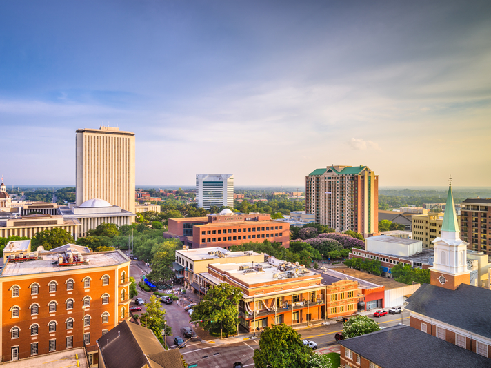 Aerial view of a cityscape with mid-rise and high-rise buildings, tree-lined streets, and a church steeple under a clear blue sky in the early evening.
