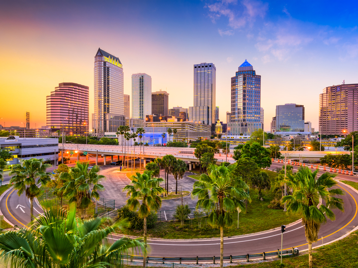 Sunset view of a downtown city skyline with tall buildings, palm trees, and a curved highway in the foreground under a colorful sky.