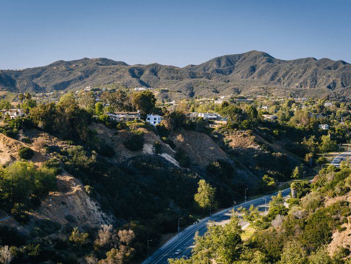 A scenic view of winding roads and houses nestled among green hills, with mountains in the background under a clear blue sky.