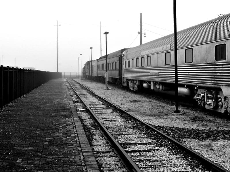 A black-and-white photo of a deserted train station platform with empty tracks beside stationary vintage train cars on a foggy day. No people are visible; the scene is quiet and still.