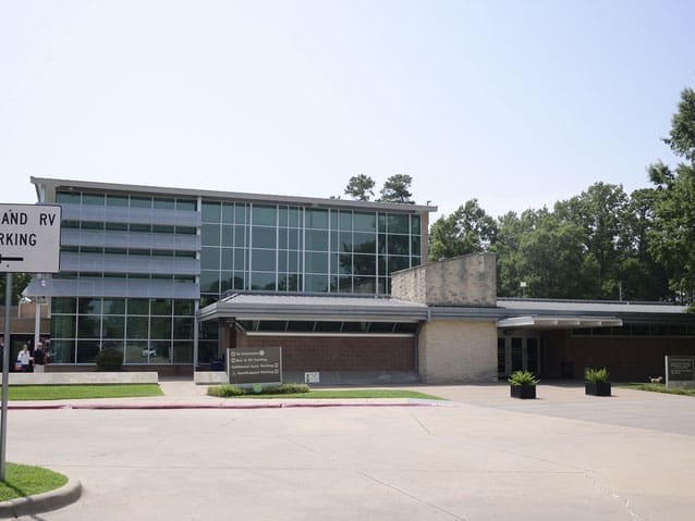 A modern building with large glass windows and brick walls, set behind a wide driveway and surrounded by trees. A street sign and two rectangular planters are visible in front of the entrance.