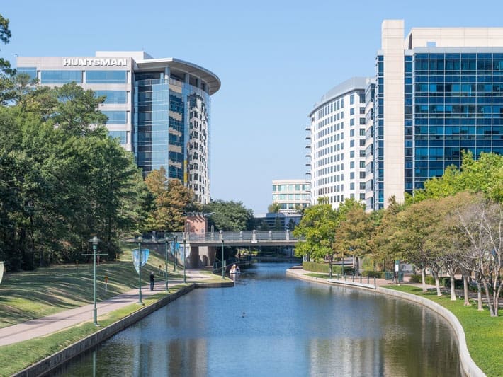 A canal runs between two modern office buildings with glass facades, surrounded by trees and a walking path on a sunny day. One building has a HUNTSMAN sign on top.