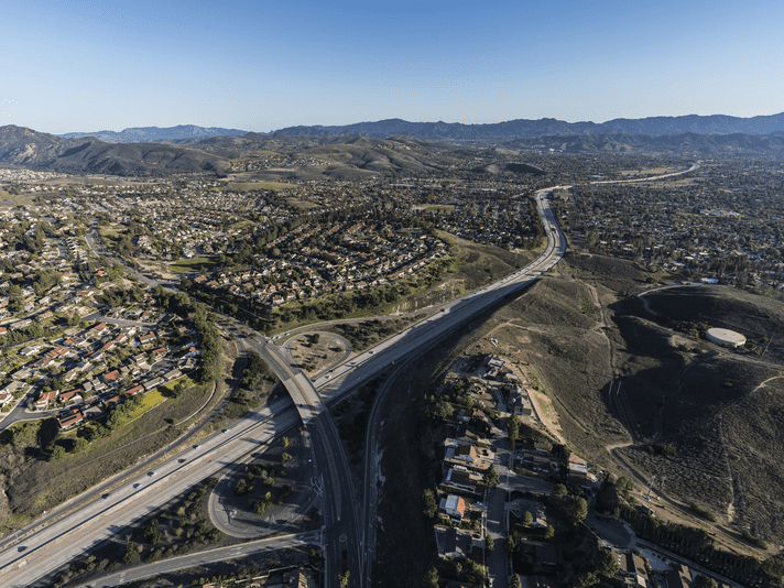 Aerial view of a suburban area with winding roads, houses, and green hills under a clear blue sky, with mountains visible in the distance.