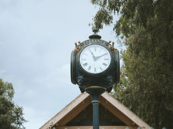 A vintage-style black clock labeled “Torrance” stands outdoors atop a post, with Roman numerals on its face and trees and a peaked roof in the background. The time on the clock is 10:12.