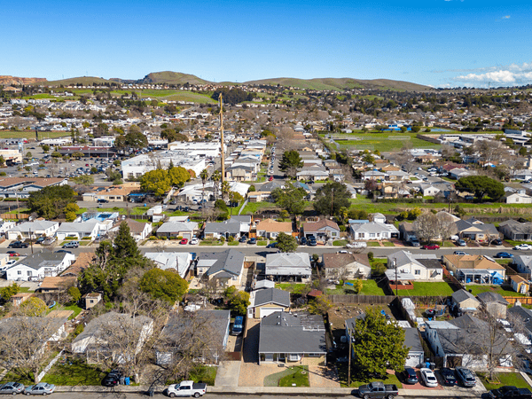 Aerial view of a suburban neighborhood with rows of houses, green lawns, and tree-lined streets, set against rolling hills and a clear blue sky in the background.