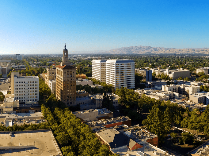 Aerial view of downtown San Jose, California, featuring mid-rise office buildings, a prominent clock tower, tree-lined streets, and distant hills under a clear blue sky.