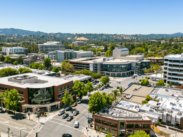 Aerial view of a suburban downtown area with low-rise buildings, tree-lined streets, parked cars, and hills in the background on a clear sunny day.