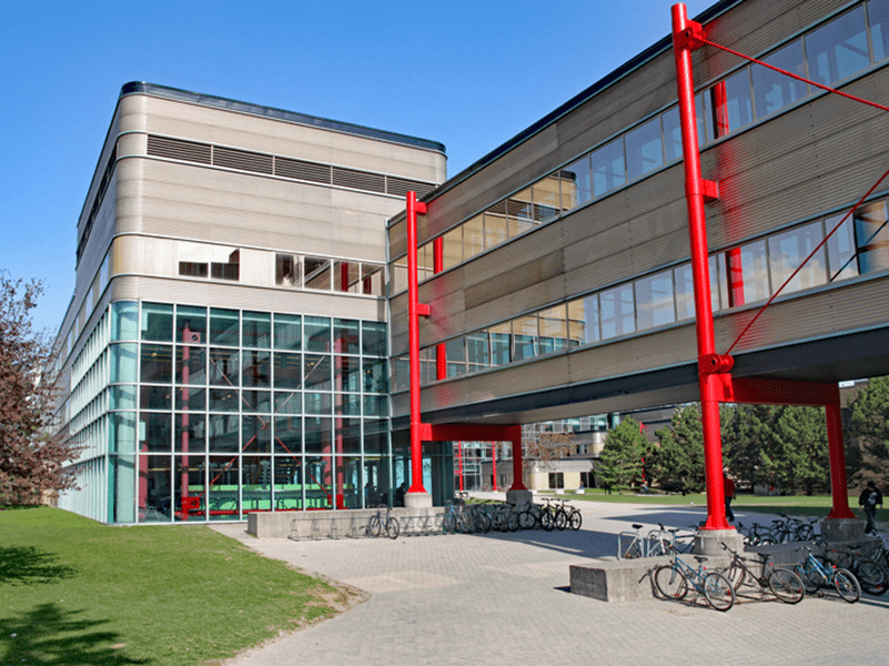 A modern building with large glass windows and a covered walkway supported by bright red columns, surrounded by green grass and several parked bicycles on a sunny day.