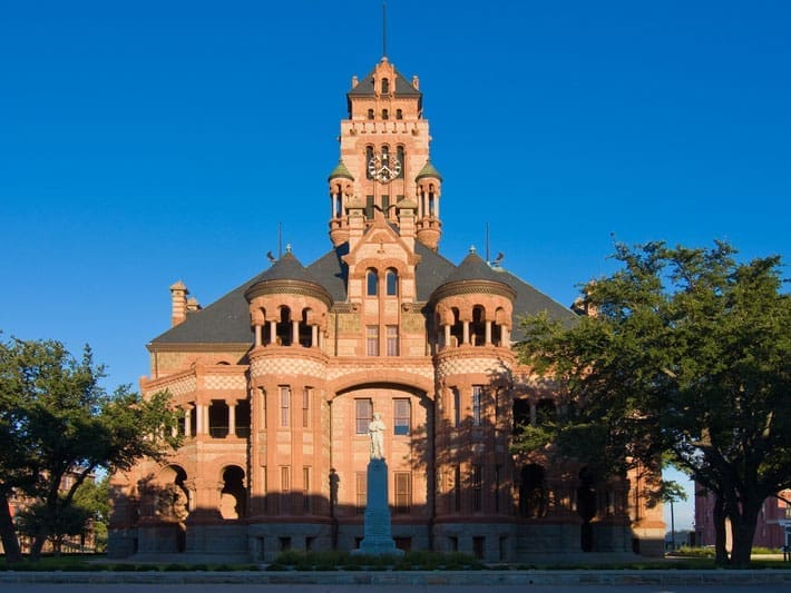 A historic red brick courthouse with a central clock tower, arched windows, and decorative details, framed by green trees and a clear blue sky. A white statue stands in front of the building.